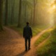 Mann steht an Weggabelung im Wald, Sonnenlicht fällt auf den naturbelassenen Pfad – Symbol für Entscheidung und Selbstverantwortung auf dem Weg zur Gesundheit